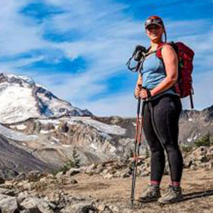Claire Nitsche stands on the top of a mountain. She carries a backpack, holds hiking sticks, and wears a cap, sleeveless top, and yoga pants.