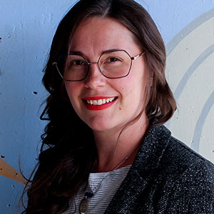 Dr. Sarah Wiebe is smiling warmly. She has brown eyes, long brown hair and white skin. She wears red lipstick, glasses, and a grey/black wool jacket.