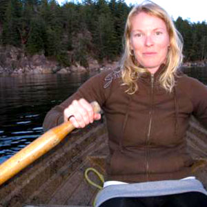 Romney McPhee is a white woman with long blonde hair. She wears a brown sweat jacket and is rowing a wooden boat. Evergreen trees are in the background.