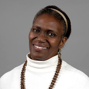 Katrina Bledsoe is a black woman with short black hair. She smiles warmly and wears a white turtleneck, necklace of brown beads, and pendant earrings.