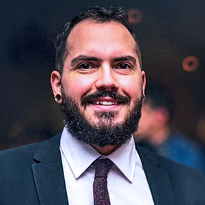 Christopher DesRivieres smiles warmly. He has black hair, light brown skin, and a full beard. He has tunnel ear piercings and wears a suit and tie.