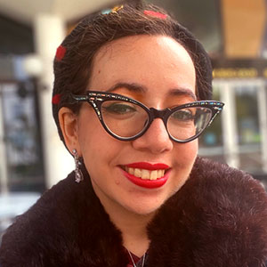 Jennifer Kaplan is a light-skinned female with a broad smile, bright red lipstick, and large cat-eye glasses. She wears a coat with faux fur collar.