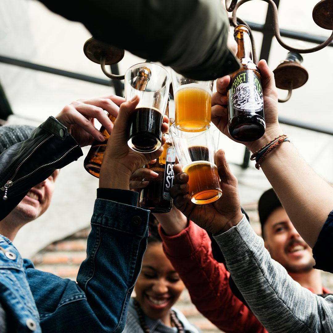 A racially diverse group of young women and men raise their glasses of beer to toast each other. They are smiling and dressed casually.