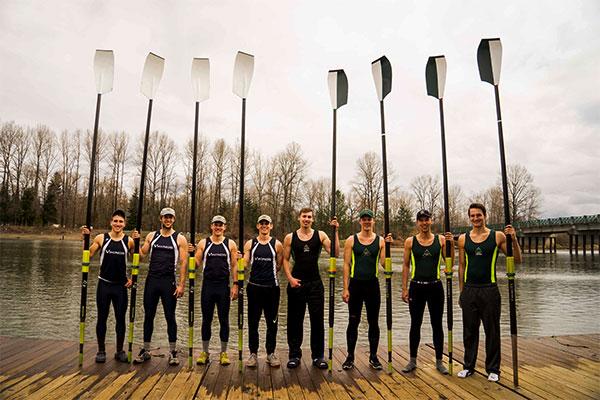 Eight male members of the WWU crew team stand on a dock holding their oars.