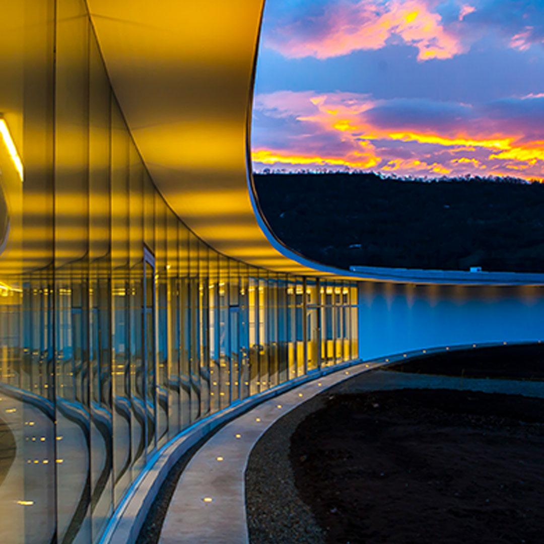 Clouds and the golden light of dusk reflect dramatically on the curved glass surfaces of a modern building.