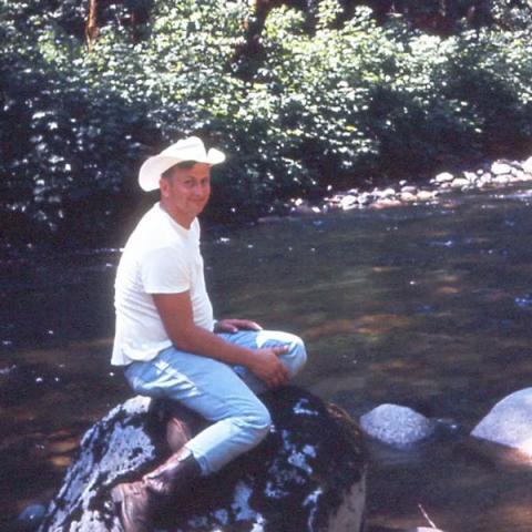 a man smiles, wearing a cowboy hat, white T, jeans and boots, sitting on a rock next to a stream.