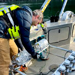 Andrew Spanjer is on a boat and bends down to inspect water monitoring equipment. He wears a warm jacket and life vest.