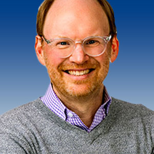 Mark Staton smiles broadly and is wearing a grey sweater and lavender shirt. He has white skin, short facial hair, and clear-framed glasses.