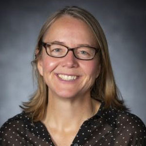 Ruth Sofield is smiling. She has white skin, blond hair, and wears glasses and a brown shirt with beige polka dots.