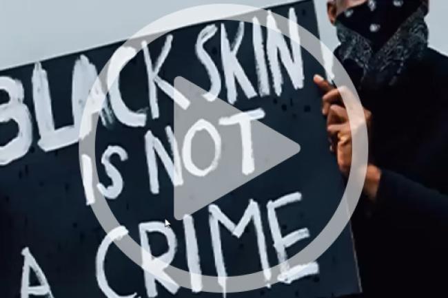 African American holding a sign that says "Black Skin is Not a Crime" with a video play button over it