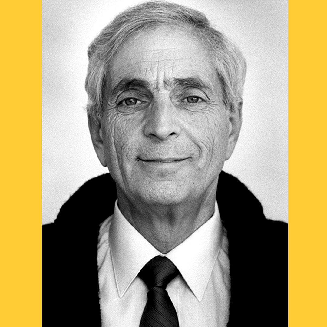 Black and white portrait of Irwin Slesnick with a yellow background. He has grey hair, bushy eyebrows, wrinkles, and a wry smile.