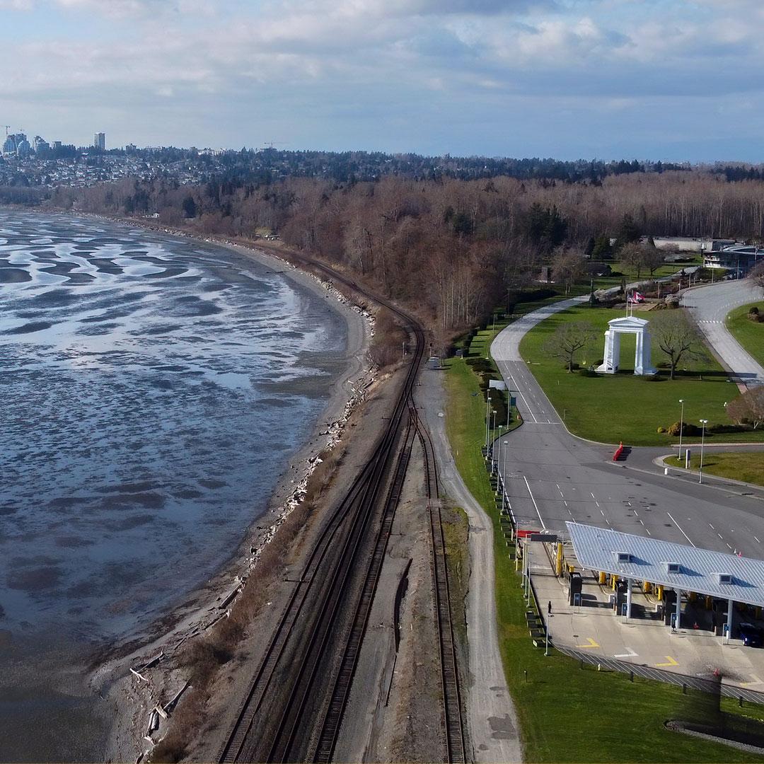Sweeping view of US/Canada border crossing at Blaine, Washington with rippling water, train tracks, trees, green grass, and the Peace Arch monument.