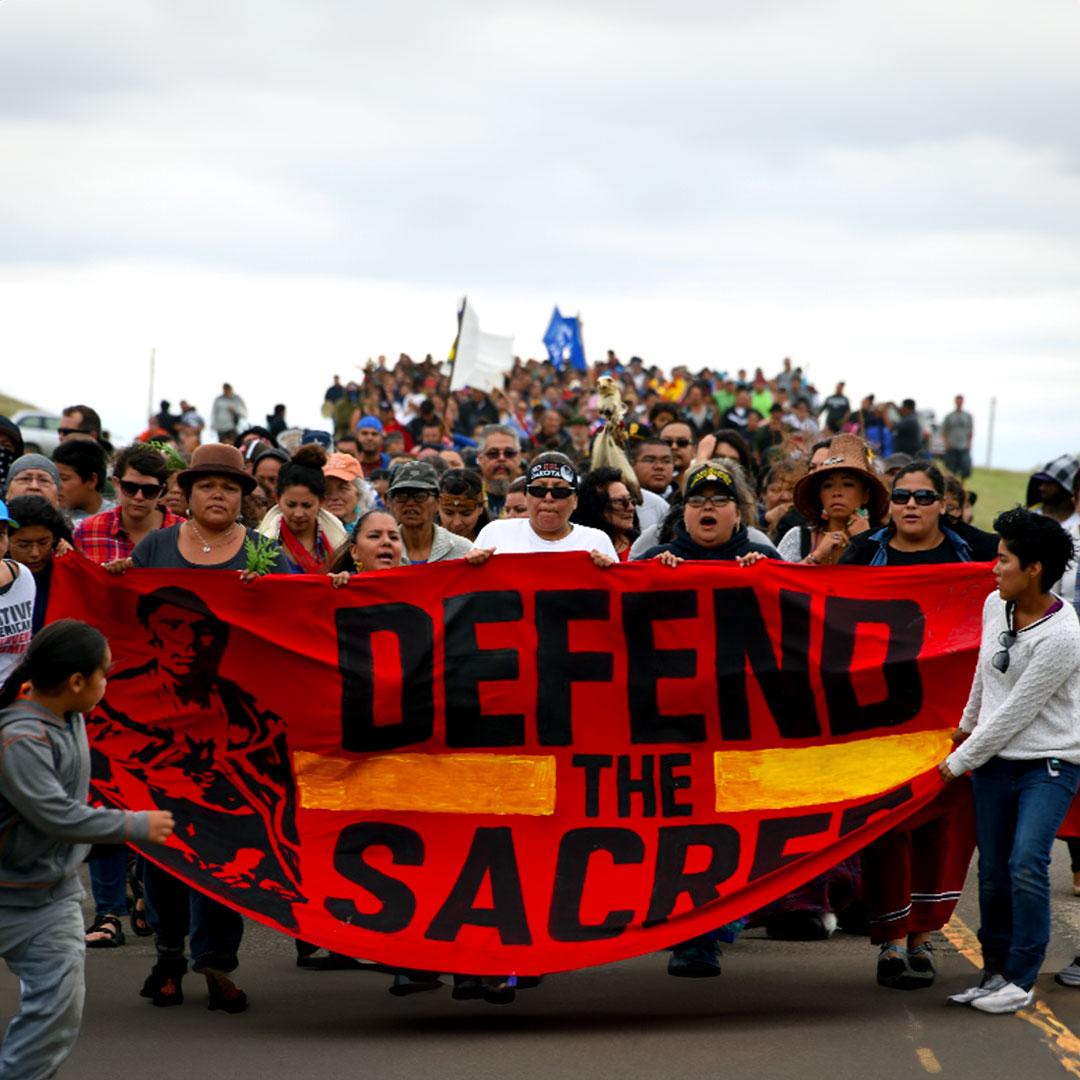 At a large outdoor march a group of Native Americans carries a large red banner that says "Defend the Sacred."